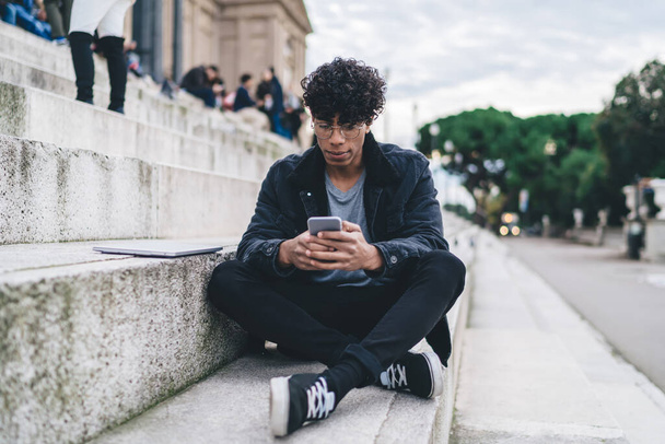 Young African American man in stylish clothing with curly hair using smartphone while sitting on stairs on blurred background outdoors - Foto, Bild