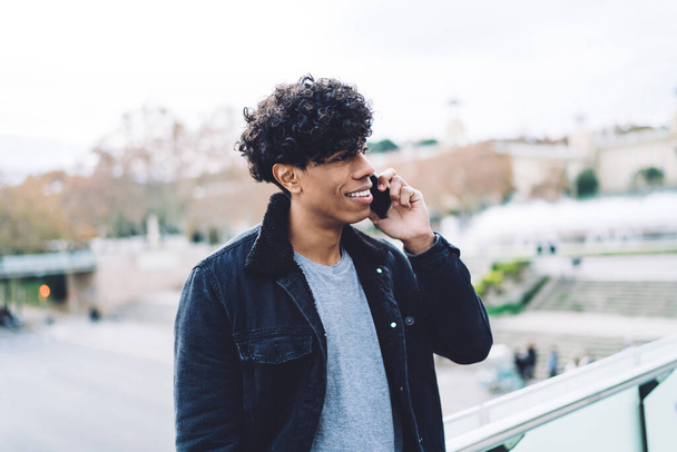 Smiling young handsome African American man with curly hair in stylish clothes talking on phone while standing on blurred background - Foto, Imagem