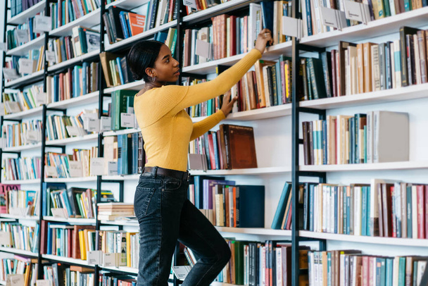Side view of young African American woman in casual outfit taking textbook from bookcase while studying in library of modern university - Fotografie, Obrázek