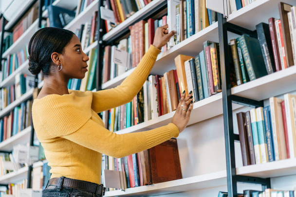 Низкий угол умной афроамериканской женщины в повседневной одежде берет учебник с полки во время проведения исследований в университетской библиотеке - Фото, изображение