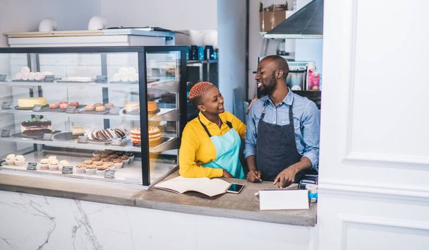 Successful cheerful African American coworkers standing at counter next to cake display fridge and talking while working together in small confectionery shop - Foto, immagini