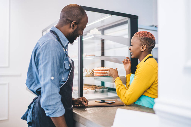 Side view of young African American owners of small confectionery shop looking at cake display fridge and discussing product line while starting new business - Fotó, kép