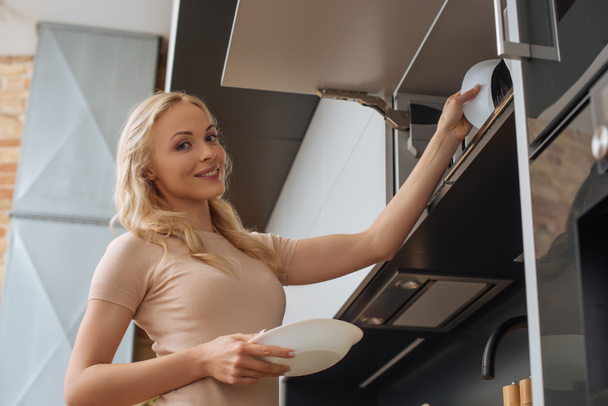 low angle view of smiling housewife putting clean plates on shelf in kitchen - Zdjęcie, obraz