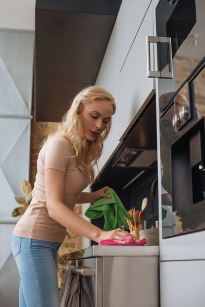 mladá hospodyňka drží gumové rukavice při otírání kuchyňského povrchu hadrem - Fotografie, Obrázek