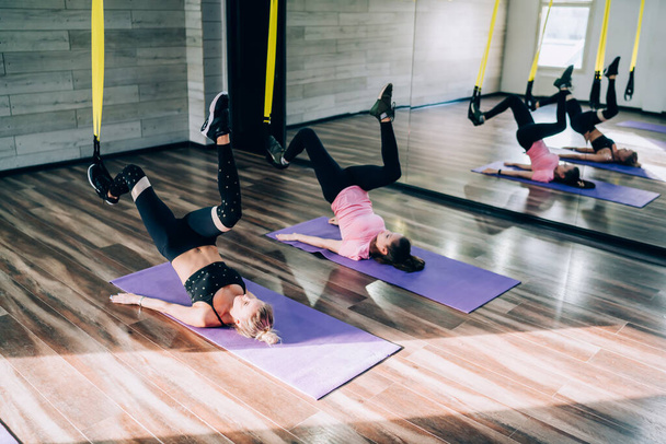 Mesdames sportives allongées sur un tapis de gym bleu et faisant de l'exercice en levant les jambes pendant l'entraînement en suspension en studio en cours de fitness - Photo, image