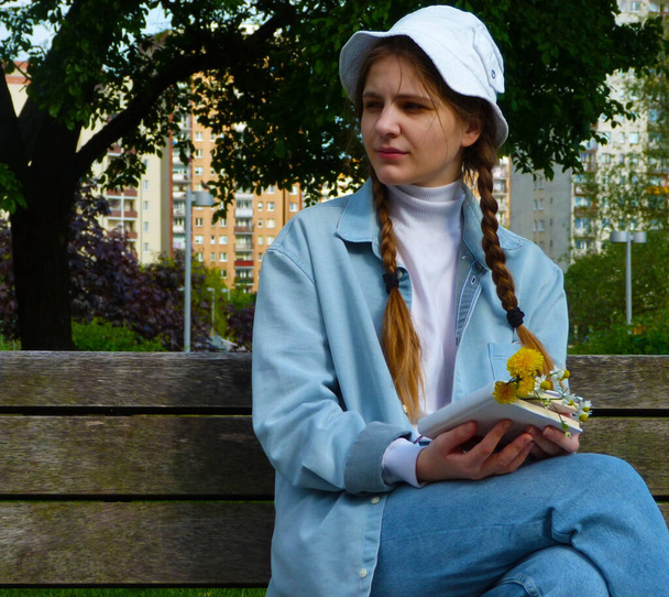 beautiful girl in a denim suit and a white panama hat sits on a bench with a book and dandelion flowers in her hands, with a high-rise building in the background - Foto, afbeelding