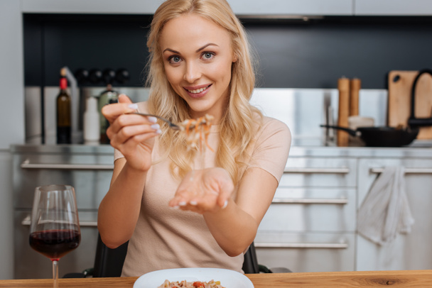 cheerful woman holding fork with thai noodles while smiling at camera near glass of red wine - Zdjęcie, obraz