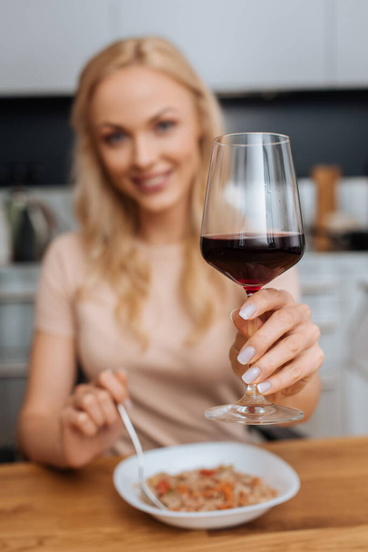 επιλεκτική εστίαση χαρούμενη γυναίκα κρατώντας ένα ποτήρι κόκκινο κρασί, ενώ κρατώντας πιρούνι κοντά στο πιάτο με thai noodles - Φωτογραφία, εικόνα