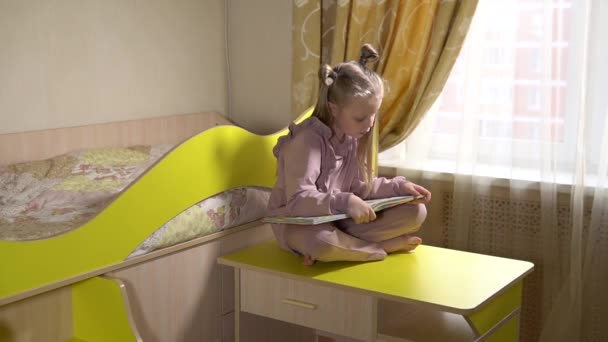 Happy reads a book sitting on the stairs on wheels with a joyful face little girl in the childrens room in fashionable clothes. On against a sunny window. - Кадри, відео