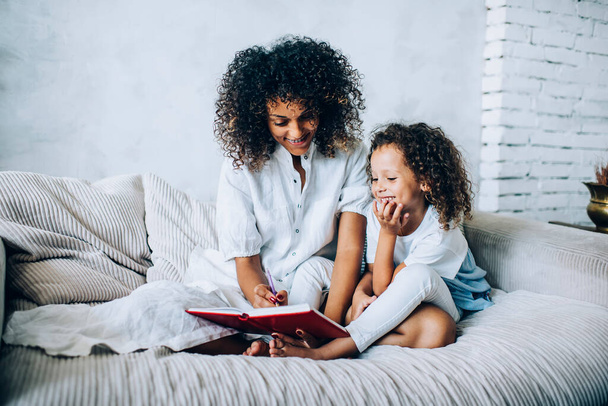 Happy adult ethnic mother in white clothes sitting on sofa with charming girl and reading book in room at home - Foto, Imagem