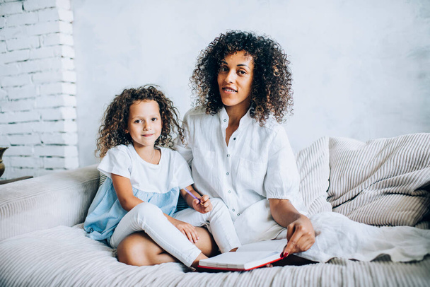 Happy curly haired ethnic mother in white clothes and charming little daughter sitting on cozy gray sofa with book smiling at camera - Foto, Bild