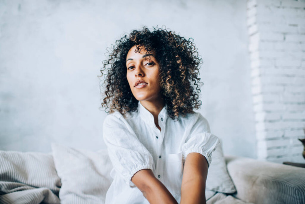 Peaceful focused ethnic lady with Afro hairstyle in white blouse chilling alone on cozy couch in apartment looking at camera - Фото, изображение