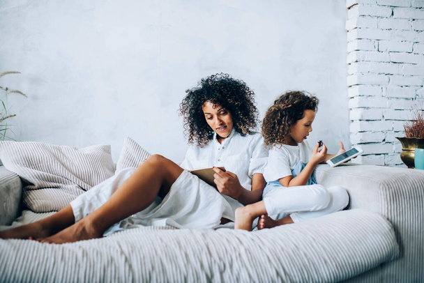 Adult ethnic woman reading book while sitting on cozy sofa with little girl surfing mobile phone and eating cookie spending time together at home - Photo, Image