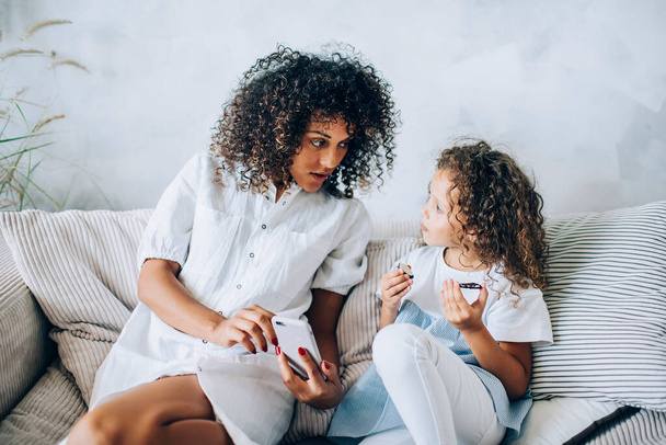 Concentrated little cute child eating biscuits and listening mom explaining educational game on smartphone while sitting together on sofa at home - Foto, Imagen