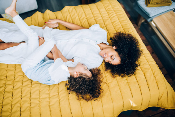 From above carefree youthful ethnic mommy and adorable little girl cuddling and looking at each other with love while laying on bed in light room - 写真・画像