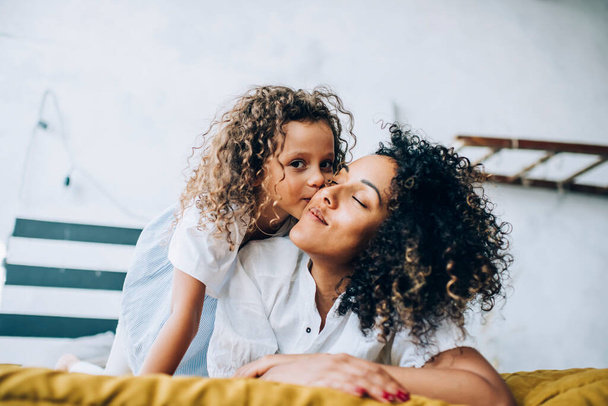 Adorable child hugging and kissing cheek of ethnic glad woman with curly hairstyle in casual wear relaxing in bedroom at home looking at camera - Foto, Imagen