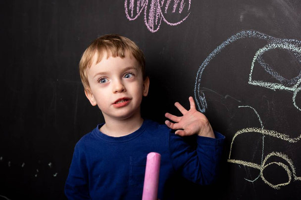 Kleine jongen leert tekenen met krijt op een zwarte leistenen muur. Kleurrijke tekeningen voor kinderen. tekenlessen. Het kind toont zijn tekening aan de muur. Europees uiterlijk. close-up. - Foto, afbeelding