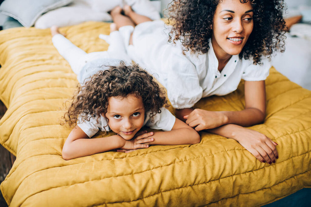 Ethnic bright curly haired mother in casual clothes relaxing on bed with dreaming girl leaning on hands at home looking at camera - Фото, изображение