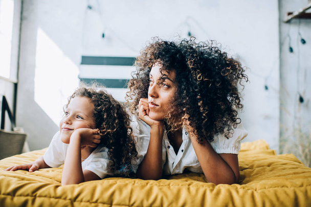 Ethnic curly haired mother in casual wear leaning on hands laying on bed with dreaming girl at home looking away - Фото, зображення