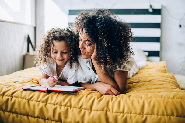 Joyful baby writing in notebook lying with curly haired mother in casual wear leaning on hand in bedroom at home - 写真・画像