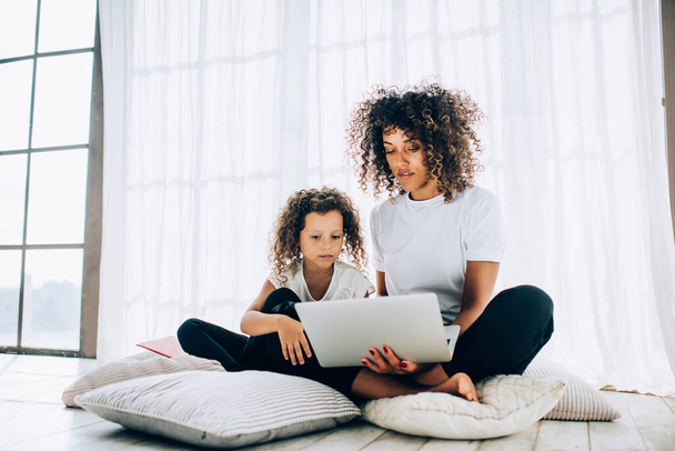 Slim ethnic mother in casual clothes watching laptop sitting with concentrated adorable curly haired daughter on pillows in light living room - Valokuva, kuva