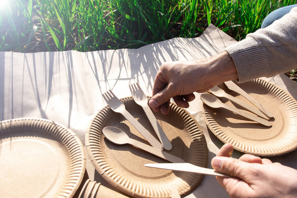 El proceso de servir vajilla desechable biodegradable en un picnic. La mano masculina distingue los tenedores de madera, las cucharas, los cuchillos en la plancha de papel por la naturaleza. hierba alrededor. respetuoso del medio ambiente
 - Foto, imagen