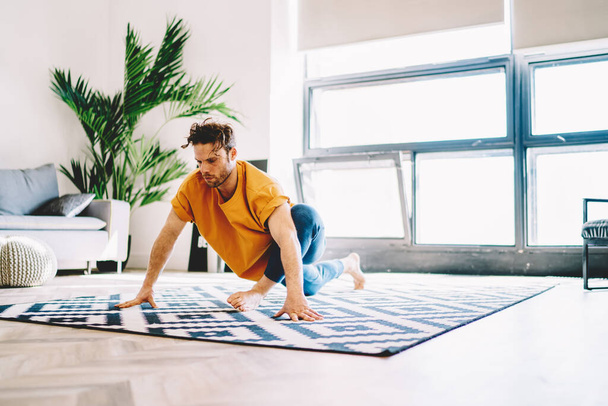 Young strong male in orange t shirt and blue pants doing yoga on carpet in room with big windows and plant in pot - Фото, зображення
