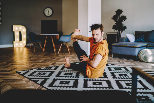 Young strong bearded male twisting and doing yoga asnaa while training at home sitting on carpet in light room - 写真・画像