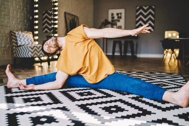Barefoot focused male in sitting on carpet in light room and bending aside stretching body in living room while doing yoga - Fotografie, Obrázek