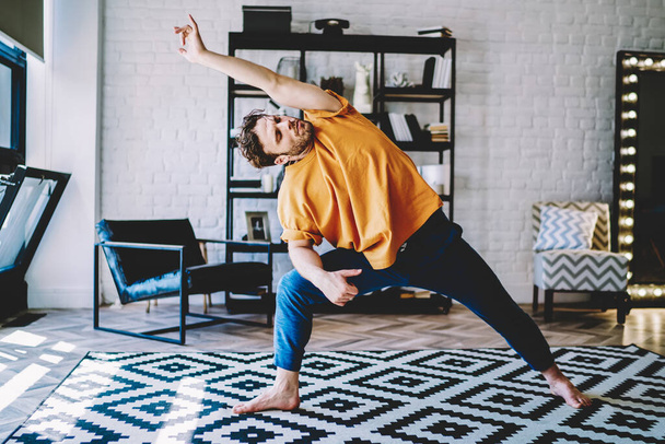 Focused athletic strong male in orange t shirt and blue pants doing yoga bending sideways on white and black carpet in room - Fotografie, Obrázek