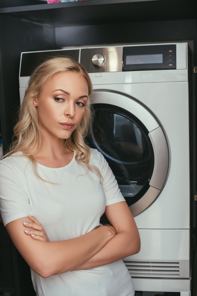 недовольная домохозяйка, стоящая со скрещенными руками возле стиральной машины
 - Фото, изображение