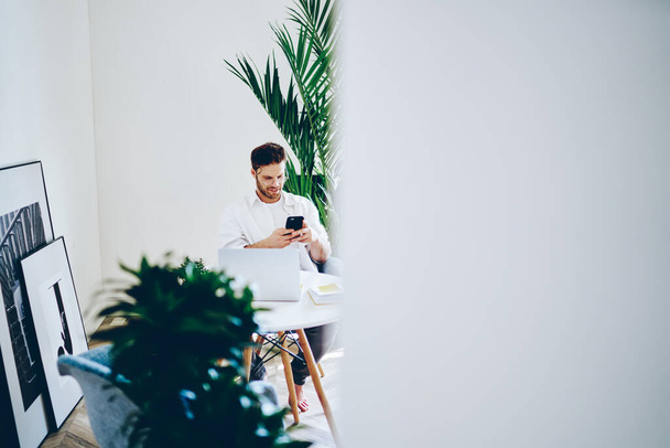 Bearded young male in white shirt and white t shirt making call on smartphone sitting at table with laptop  in creative workplace with potted plants - Foto, Bild