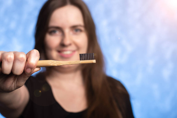 eco-friendly bamboo toothbrush in a female hand. girl with a snow-white smile on the background in defocus. oral hygiene. items from biodegradable materials. close-up. - Photo, image