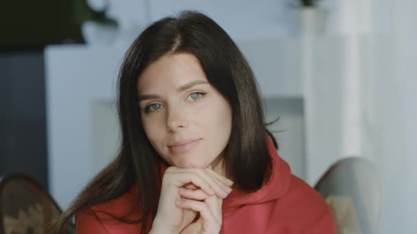 Friendly Young brunette woman in red hoodie posing for close up portrait indoors, looking at camera.  - Filmmaterial, Video