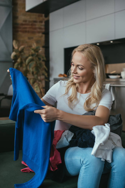 Lächelnde Hausfrau blickt auf Pullover, während sie mit Wäsche auf dem Schoß sitzt - Foto, Bild
