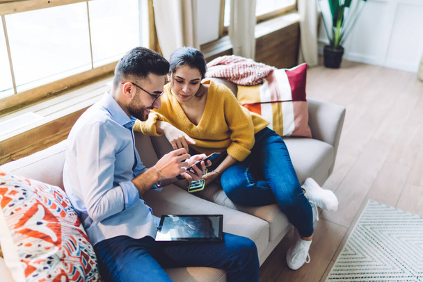 Cheerful young couple wearing jeans sitting on couch at home with tablet and sharing smartphone while spending time together at home - Foto, Imagen