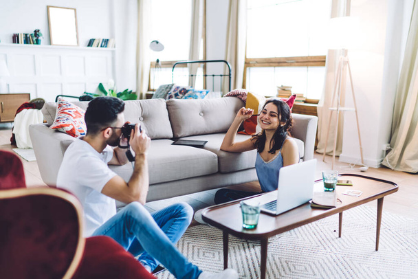 Young couple sitting on floor between sofa and coffee table with laptop while man holding photo camera and taking picture of smiling woman - Fotoğraf, Görsel