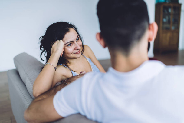 Cheerful and positive couple sitting on sofa in cozy apartment and discussing plans for next weekend while smiling and looking at each other - Photo, image
