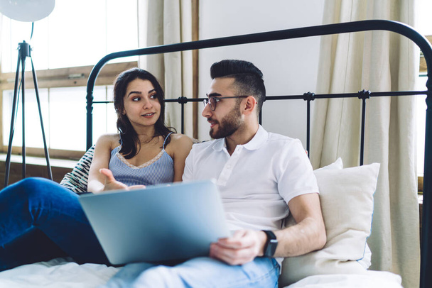 Pretty adult woman in casual clothes talking with bearded handsome man in glasses with laptop on knees while sitting on bed in light apartment - Photo, Image