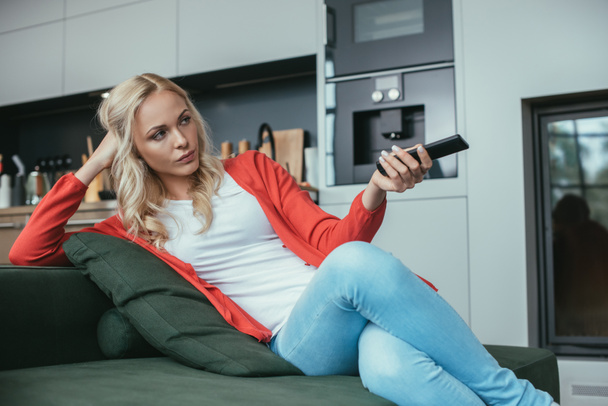 serious woman sitting on sofa and holding tv remote controller - Valokuva, kuva