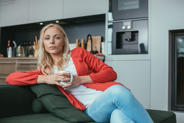 serious woman looking at camera while sitting on sofa with cup of tea - 写真・画像