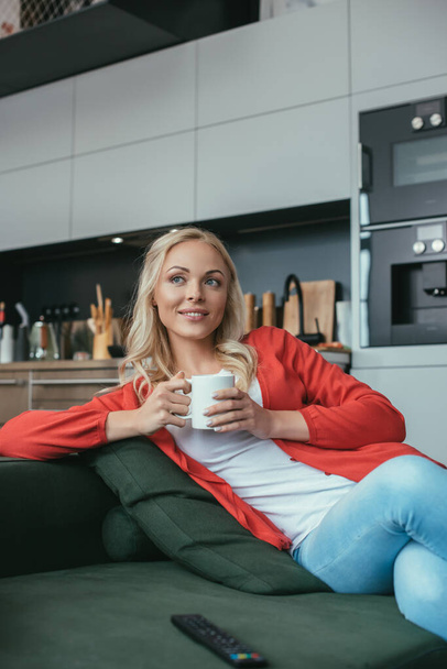 boldog nő néz félre, miközben ül a kanapén egy csésze teát - Fotó, kép