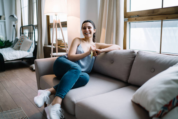 Happy young woman in tank top and jeans leaning on couch cushion and laughing while holding smartphone and looking away in apartment  - Photo, Image