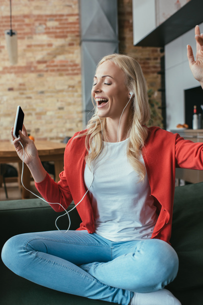 mujer feliz en auriculares cantando con los ojos cerrados y la mano levantada mientras sostiene el teléfono inteligente
 - Foto, imagen