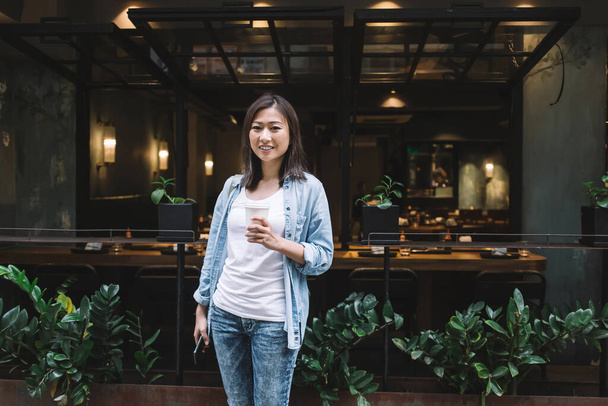 Smiling young beautiful Asian woman in casual jeans outfit browsing smartphone while holding takeaway drink standing in front of modern cafe with tropical plants looking at camera  - Foto, imagen