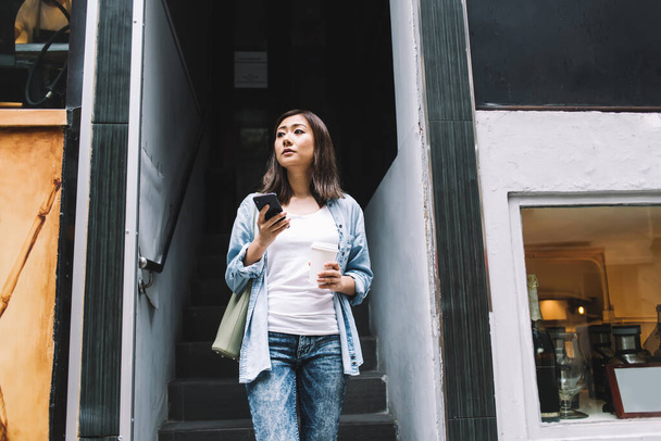 Young pretty Asian woman in casual jeans outfit holding bag browsing smartphone while holding takeaway drink standing on stairs of shop on street looking away  - Foto, imagen