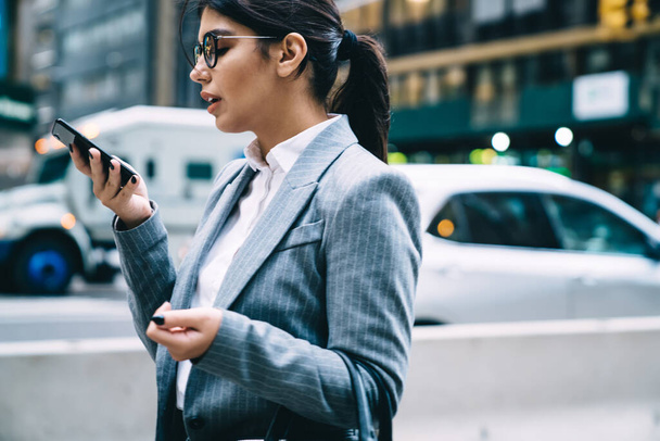 Side view of personable young female entrepreneur in glasses and formal suit standing at street with smartphone during phone conversation  - Foto, Imagem