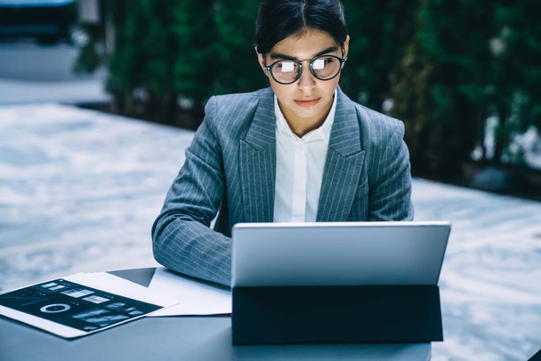 Young ethnic woman wearing office jacket and glasses sitting at tablet with paperwork working attentively on tablet looking at screen - Photo, Image