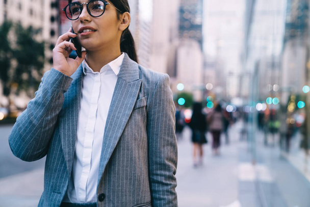 Crop elegant businesswoman in gray suit and eyeglasses talking on mobile while walking on street on background of blurry people looking away  - 写真・画像
