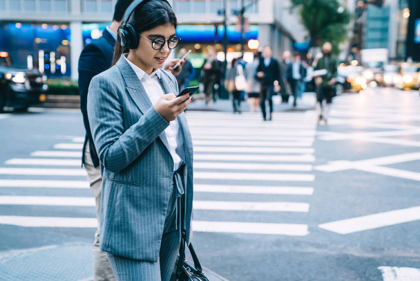 Glad female in glasses wearing office clothes browsing smartphone with interest while standing on busy city street in wireless headphones - Zdjęcie, obraz